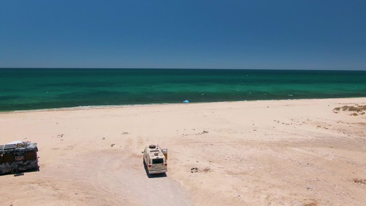 Aerial view of Playa Miramar Los Cabos with two parked vehicles separated on open sand, green-blue ocean beyond, and small surf lines near shore, with no visible people.