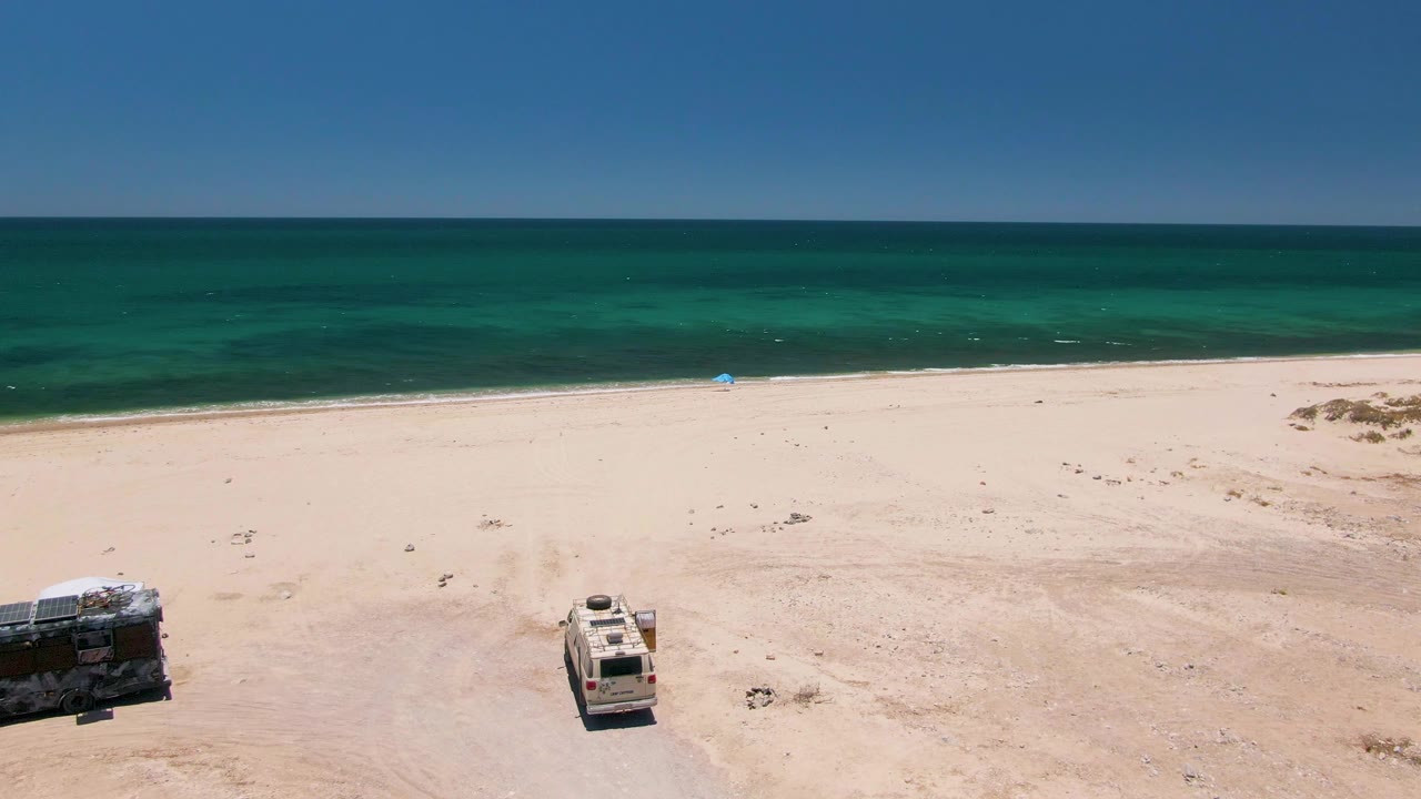 Aerial frame over Playa Miramar Los Cabos where the two vehicles sit near the lower edge of the sand and the turquoise ocean fills the background, with no visible people.