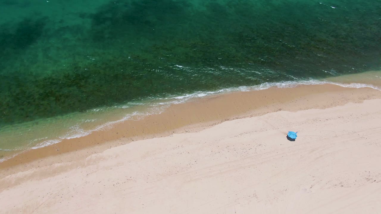 At Playa Miramar Los Cabos, turquoise water meets pale sand from above, with a small blue object on the beach and no visible people.