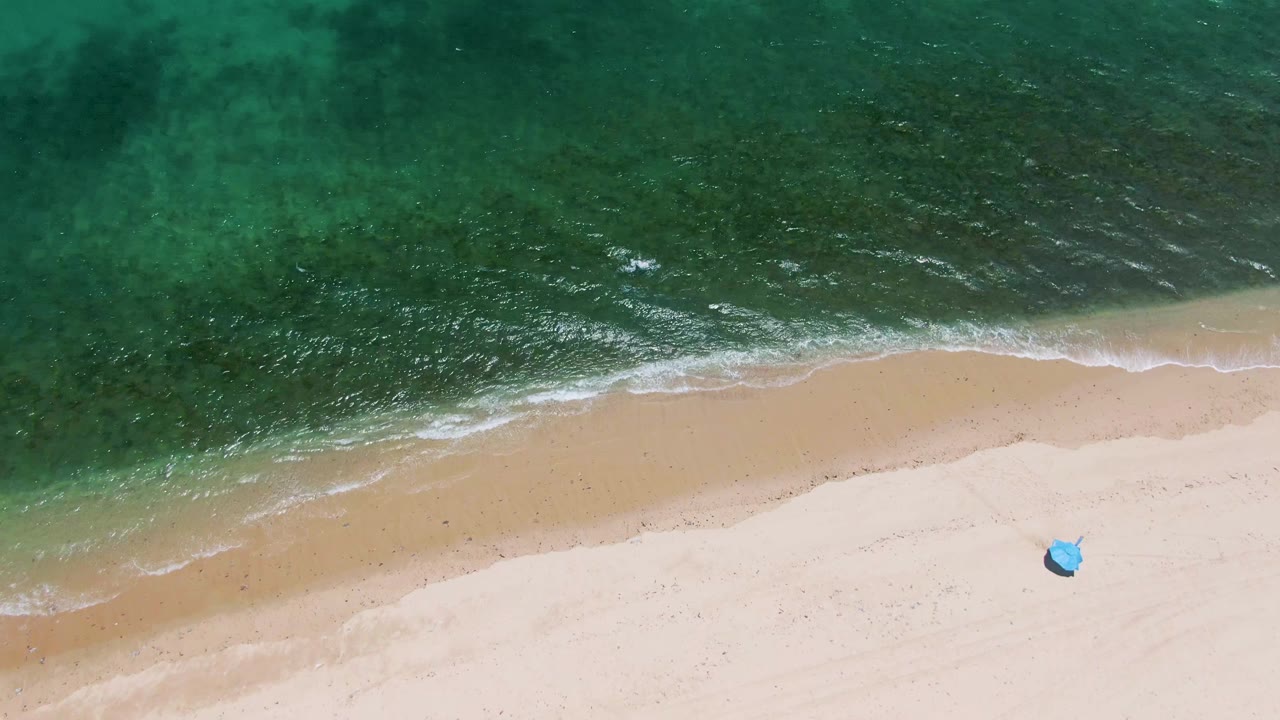 At Playa Miramar Los Cabos, clear turquoise water pushes a narrow foam line toward pale sand, with the small blue object near the beach edge and no visible people.