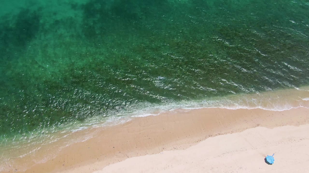 Overhead beach frame with darker turquoise water, pale sand, foam, and a small blue object at the lower right.