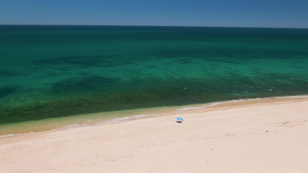 Aerial view at Playa Miramar Los Cabos with the shoreline lower in frame, turquoise water across the center, a small blue object on the sand, and no visible people.