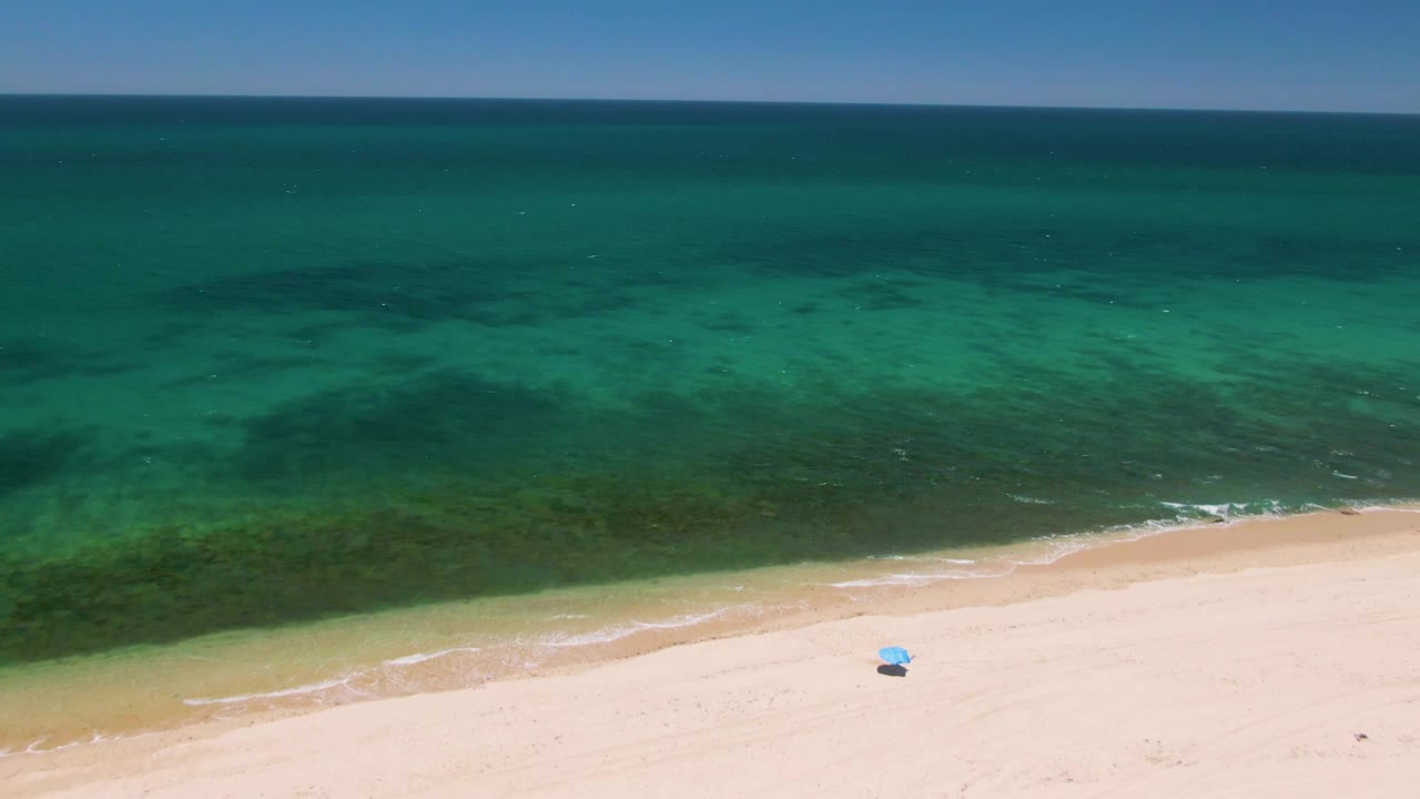 Aerial frame over Playa Miramar Los Cabos where darker green water patterns spread across the turquoise ocean above pale sand, with no visible people.