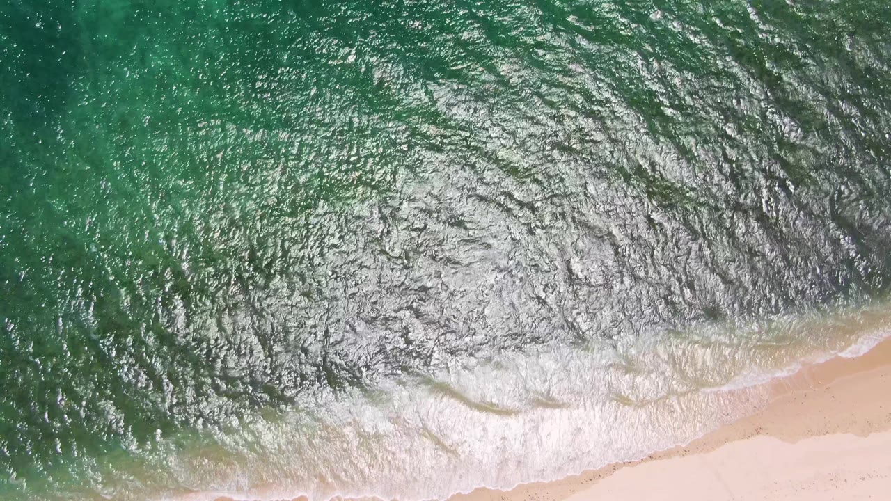 A curved white wave line crosses turquoise water near the pale beach at Playa Miramar Los Cabos, with no visible people.