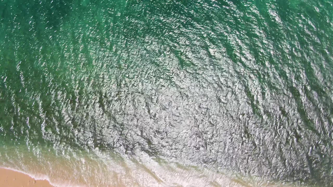 Bright sun glare spreads across rippled green water above a thin strip of sand at Playa Miramar Los Cabos, with no visible people.