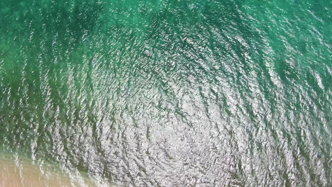 White surf spreads along the lower edge of rippled green water and pale sand at Playa Miramar Los Cabos, with no visible people.