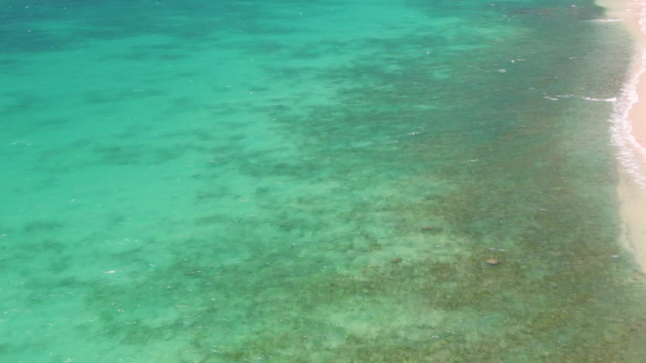 Opening aerial frame over Playa Miramar Los Cabos shows turquoise shallow water, darker submerged patches, and a narrow sandy shore on the right with no visible people.