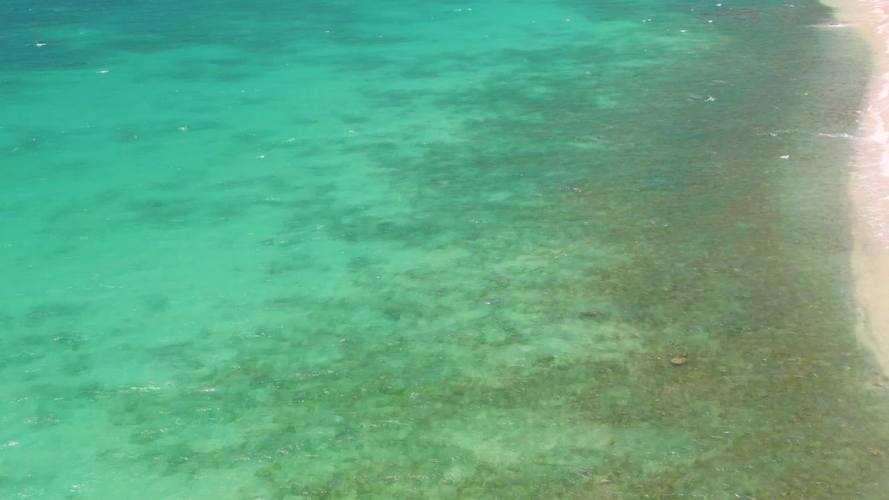 At two seconds, the aerial view shows the sandy beach strip pressed to the right side, with turquoise water and mottled darker areas across the shallows at Playa Miramar Los Cabos and no visible people.