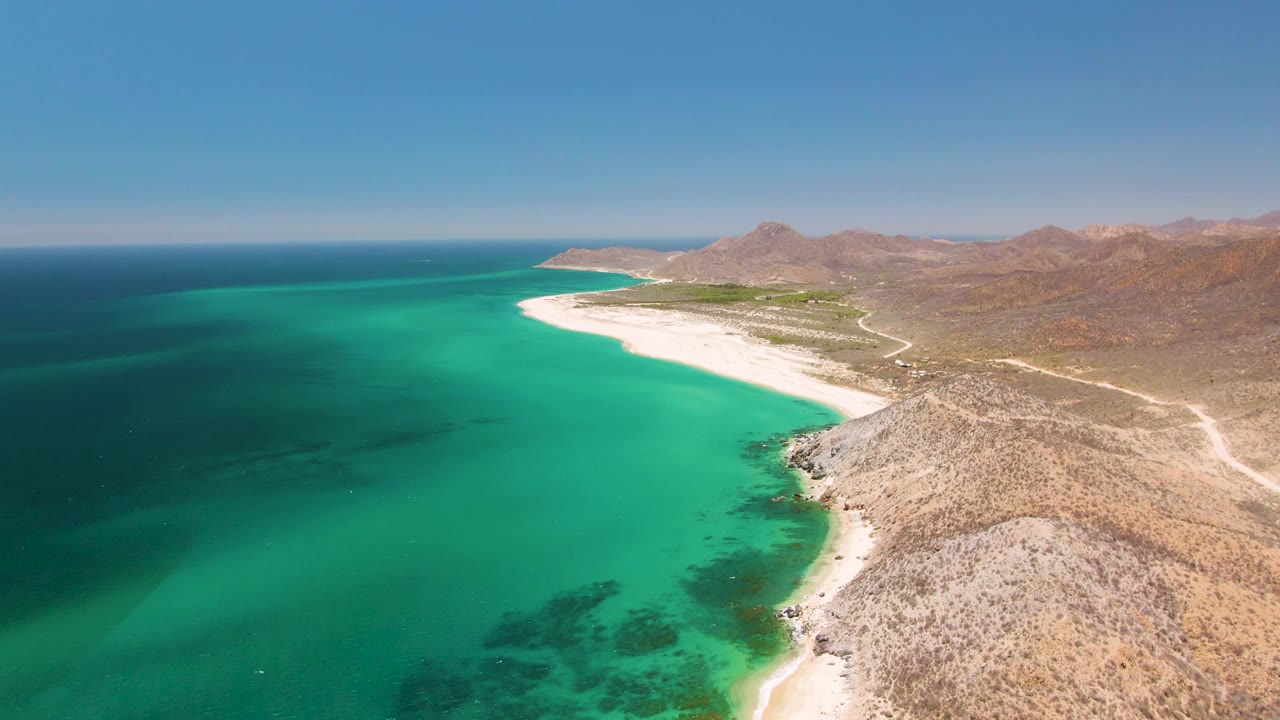 Aerial frame over Playa Miramar Los Cabos showing turquoise water beside a white sandy beach, rocky tan hills, a pale inland road, and no visible people.