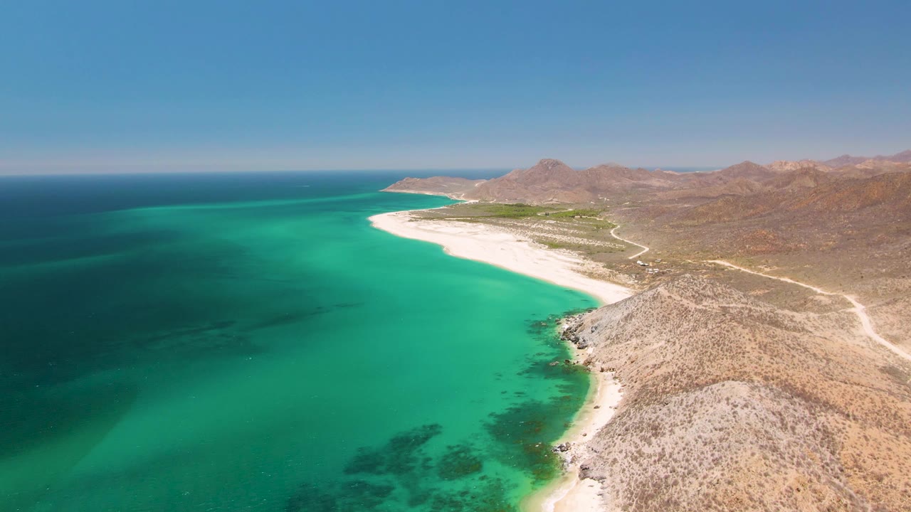 Wide drone-like frame over Playa Miramar Los Cabos showing darker offshore water, bright turquoise shallows, a rocky headland in the foreground, white sand, and no visible people.