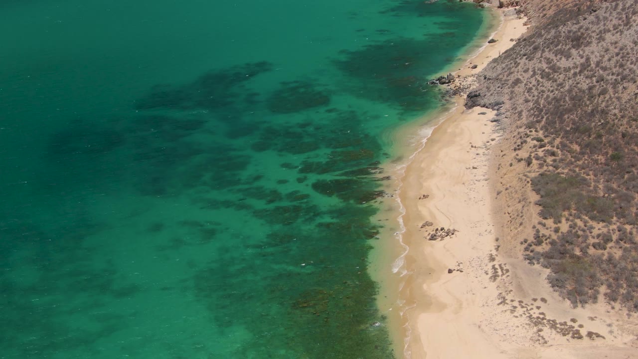 Aerial frame of Playa Miramar in Los Cabos with turquoise water on the left, pale sand near the center, dry rocky terrain on the right, and no visible people.