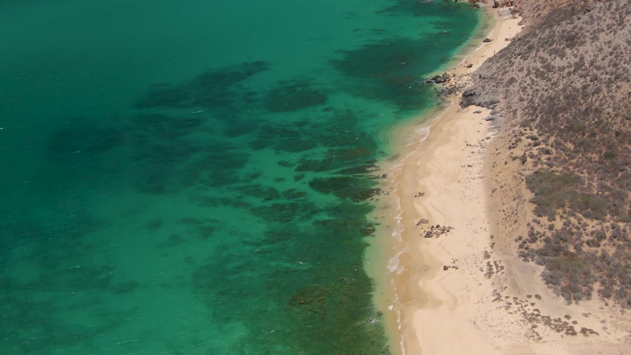 Clear green-blue ocean water curves along a sandy beach with rocky land beside it.