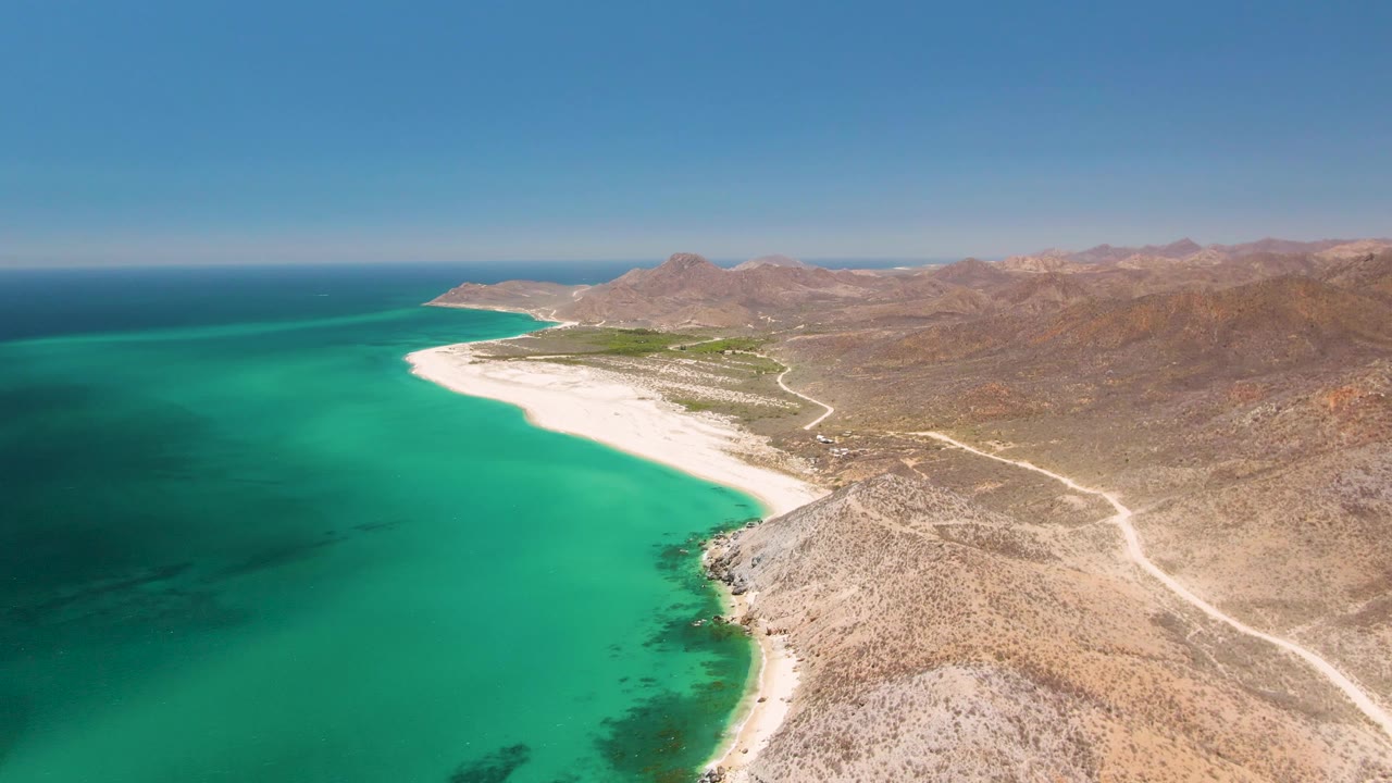 Opening aerial frame over Playa Miramar Los Cabos with turquoise ocean on the left, a curved white sand beach, rocky desert foreground, distant mountains, and no visible people.