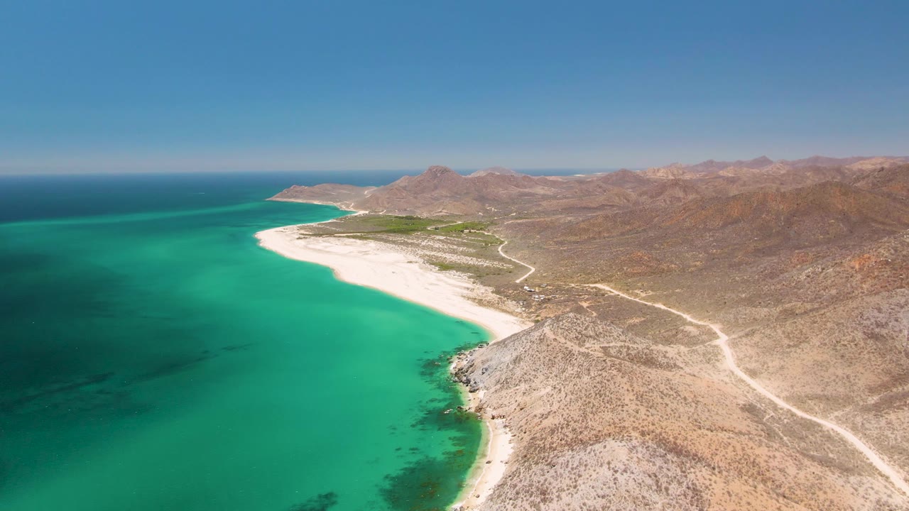 Aerial frame at Playa Miramar Los Cabos showing the beach curve more centrally, with green-blue water, pale sand, dry coastal road, brown mountains, and no visible people.