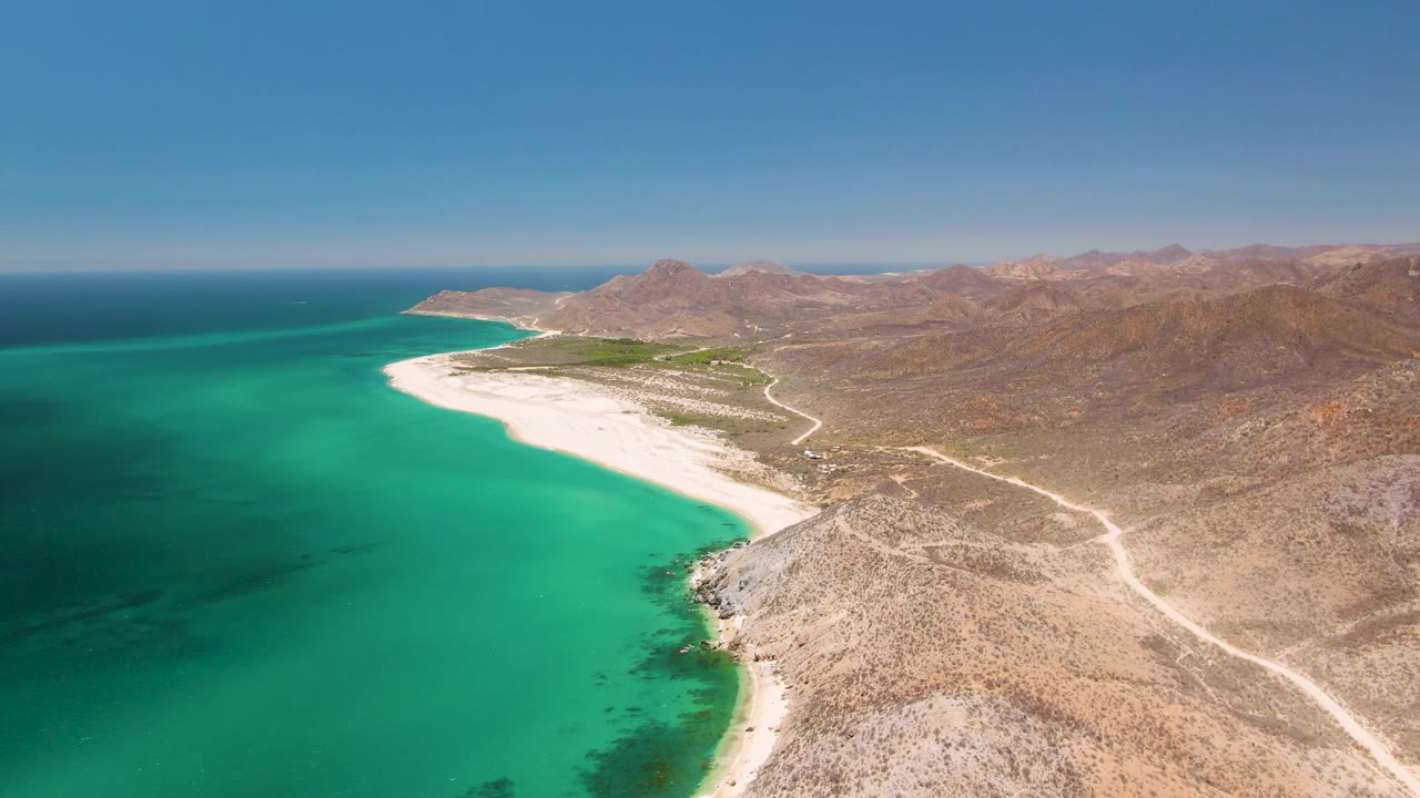 Mid-clip aerial over Playa Miramar Los Cabos with the turquoise bay filling the left side, white beach across the center, rocky desert slopes to the right, mountains beyond, and no visible people.