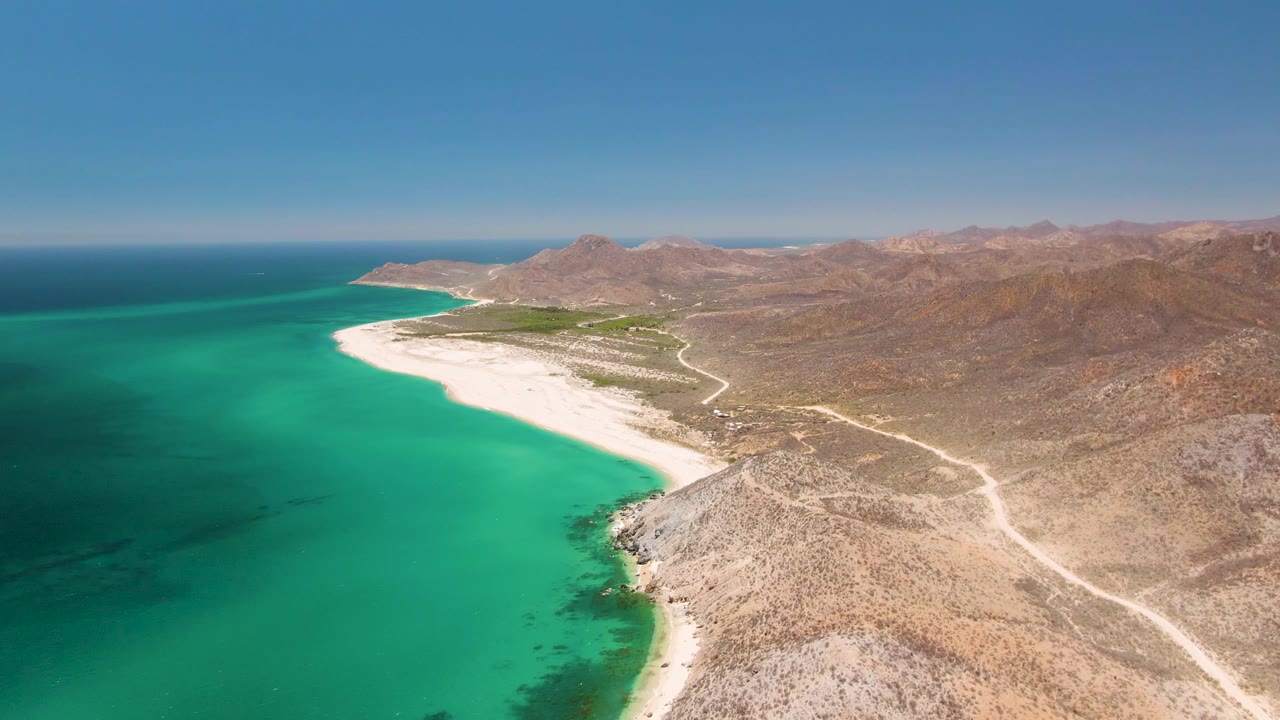 Aerial coastline with deep green-blue ocean, pale beach, dirt road, and arid mountains.