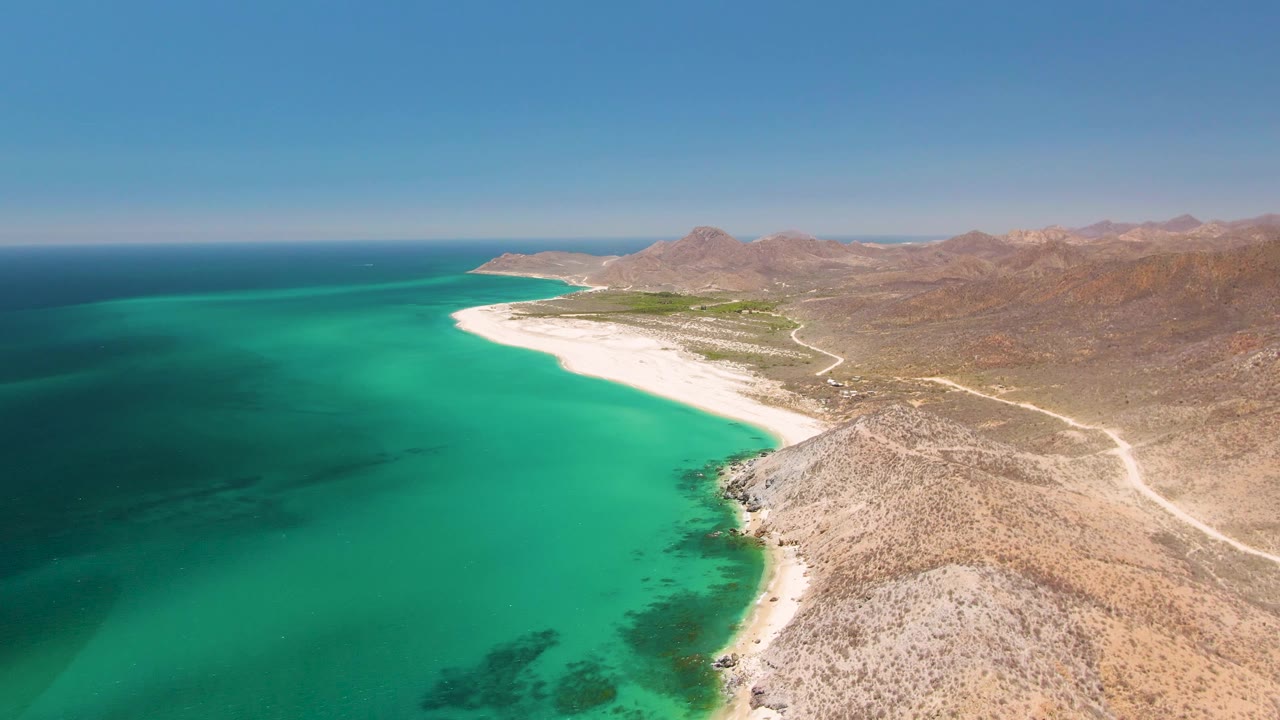 Aerial view over Playa Miramar Los Cabos with turquoise water on the left, white sand curving ahead, rocky desert shore in front, distant mountains, and no visible people.