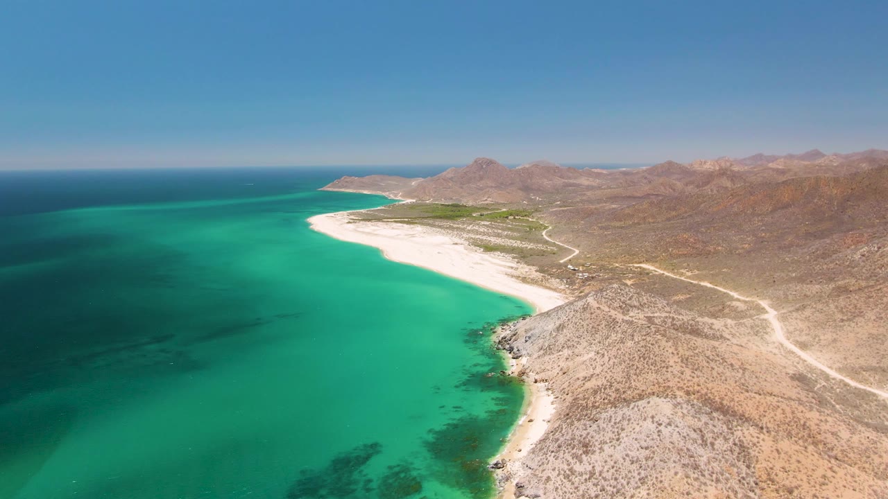High aerial frame of Playa Miramar Los Cabos showing a broad turquoise bay, pale beach, rocky point in the foreground, winding shore road, brown hills, and no visible people.