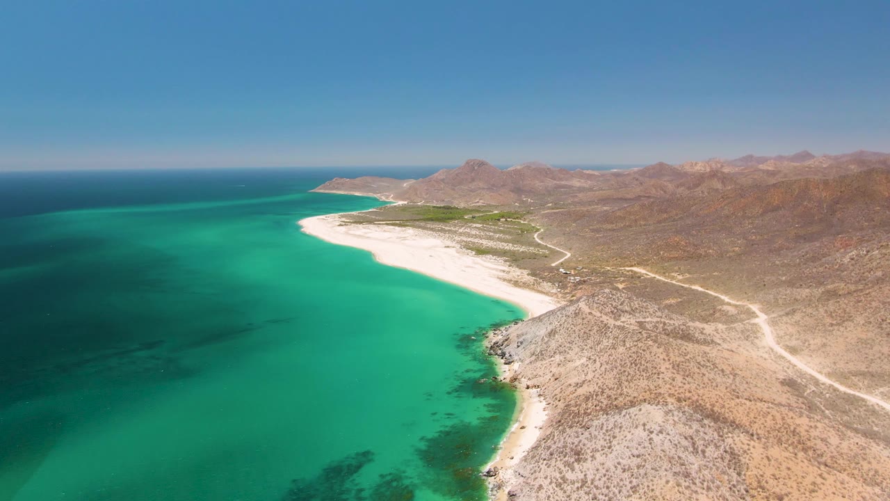 Aerial view of Playa Miramar Los Cabos with deeper blue water at far left, bright turquoise shallows near shore, white beach ahead, rocky desert slope at right, and no visible people.