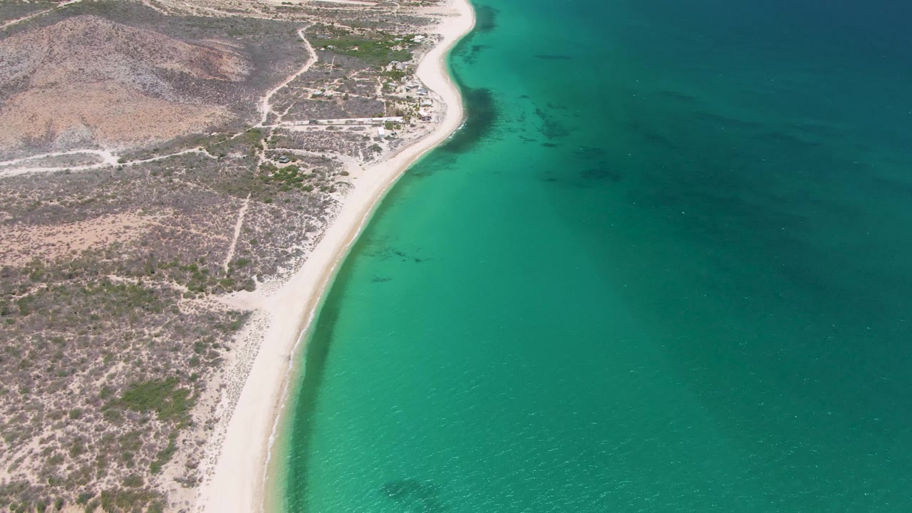 Aerial view of Playa Miramar Los Cabos with the beach running diagonally through frame, dry land on the left, clear green-blue ocean on the right, and no visible people.