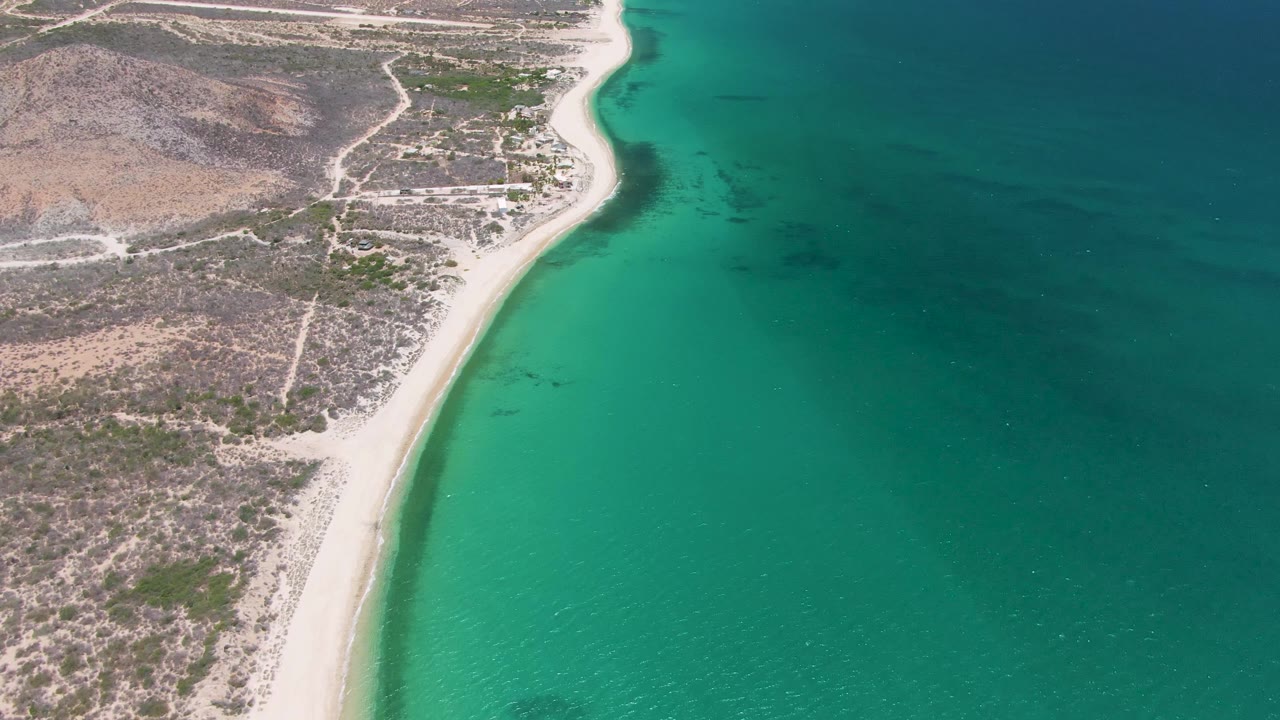 Aerial frame at Playa Miramar Los Cabos with the sandy beach along the lower center, desert scrub and tracks inland, calm turquoise water beside it, and no visible people.