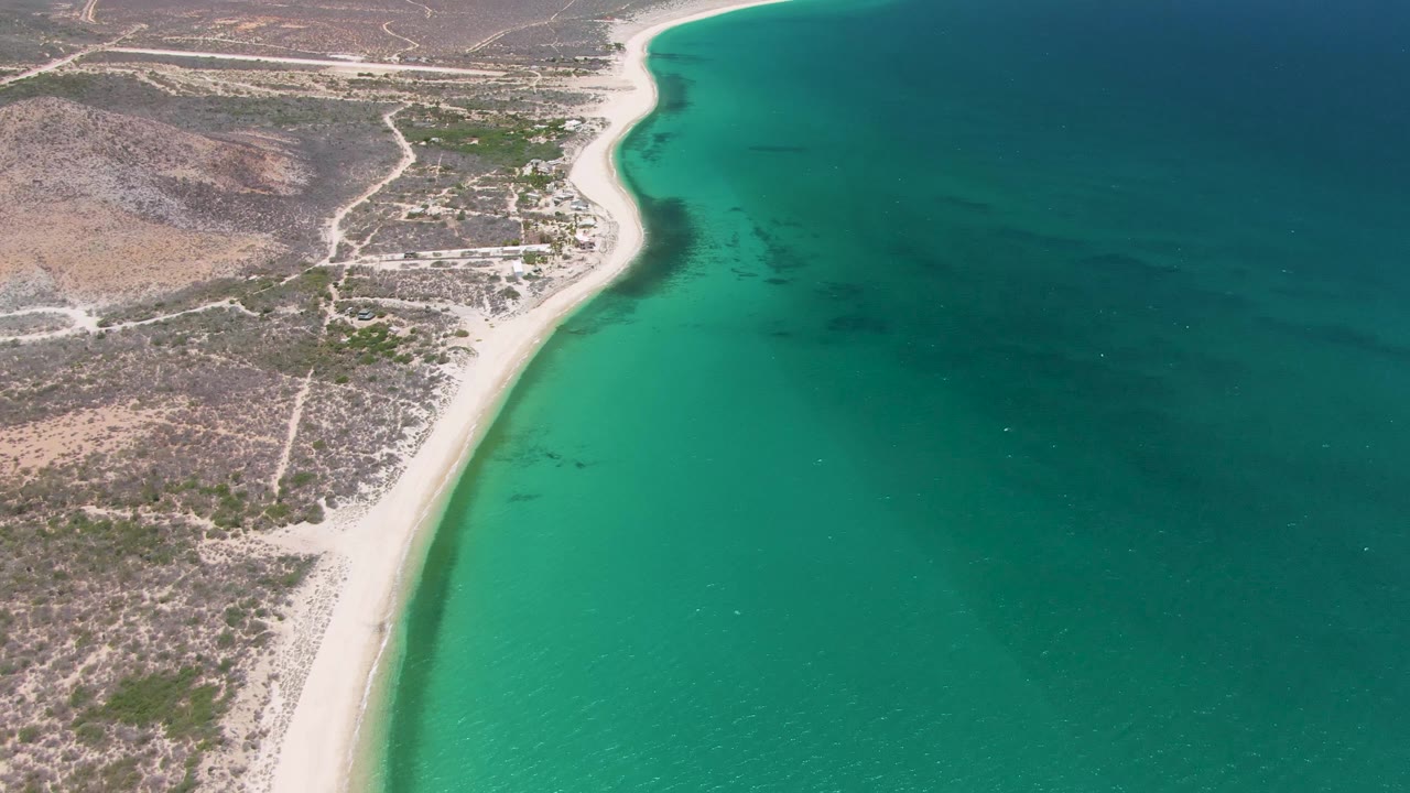 Aerial frame over Playa Miramar Los Cabos showing pale sand running along turquoise water, with dry terrain and dirt roads inland, no visible people.
