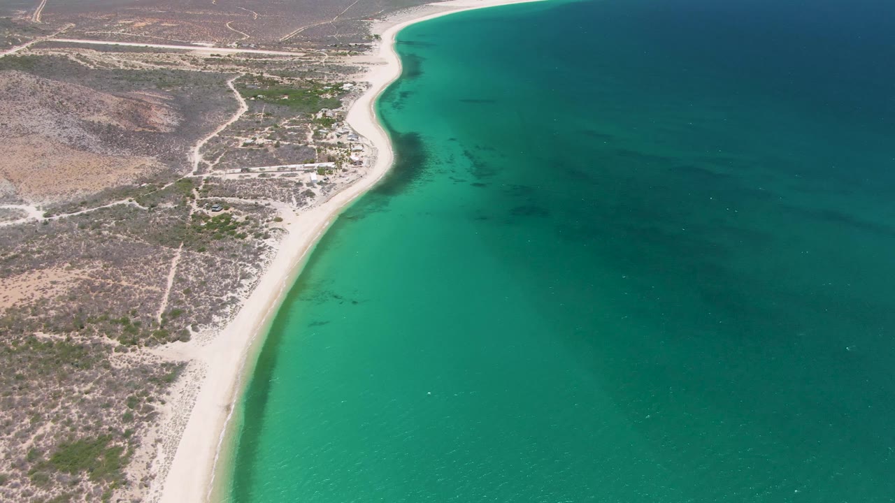 Mid-clip aerial frame of Playa Miramar Los Cabos with the narrow beach bending through the scene and open turquoise water filling much of the right side, no visible people.