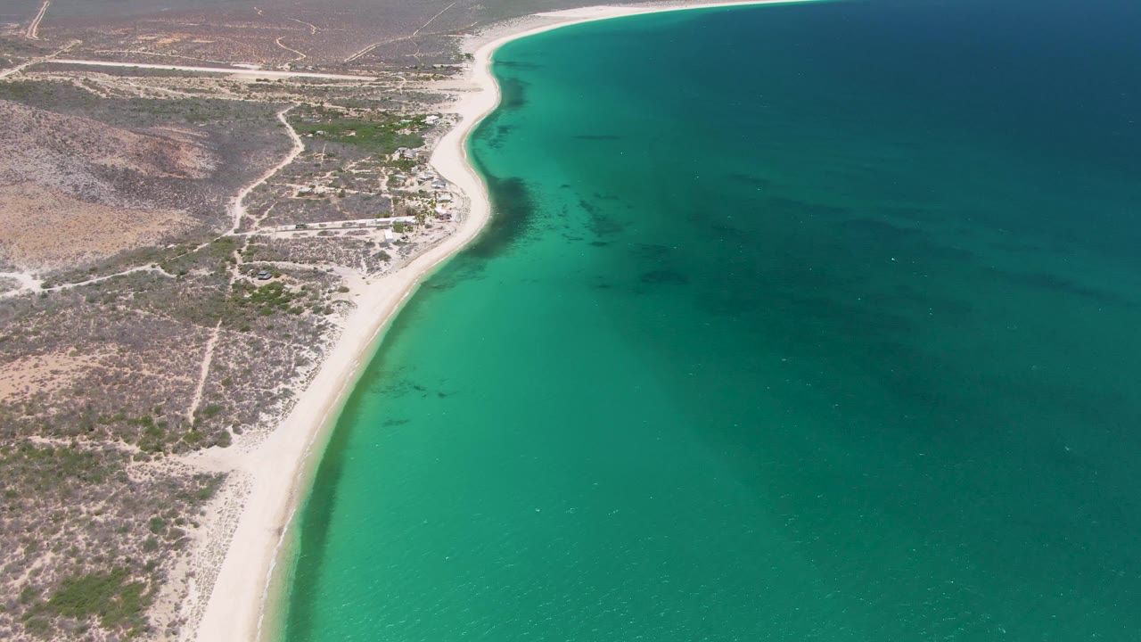 Later aerial frame over Playa Miramar Los Cabos where the curved beach remains visible along the waterline, with scrubland and roads on the inland side, no visible people.