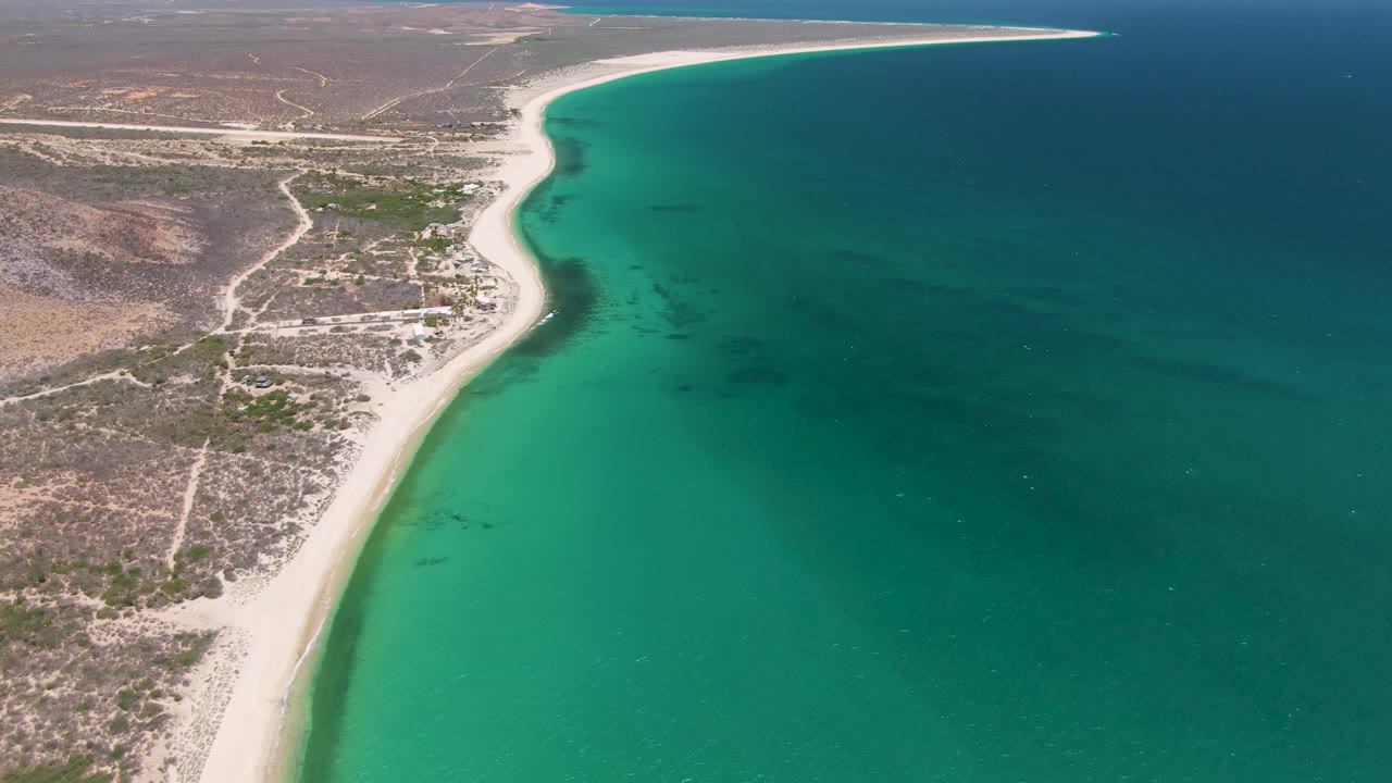 At 0:01, the Playa Miramar shoreline remains centered, with shallow green-blue water along the sand, dry inland tracks, and no visible people.