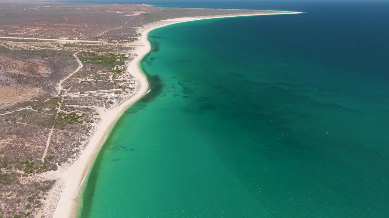 At 0:02, the aerial view emphasizes the long beach line at Playa Miramar Los Cabos, with more open ocean filling the right side and no visible people.
