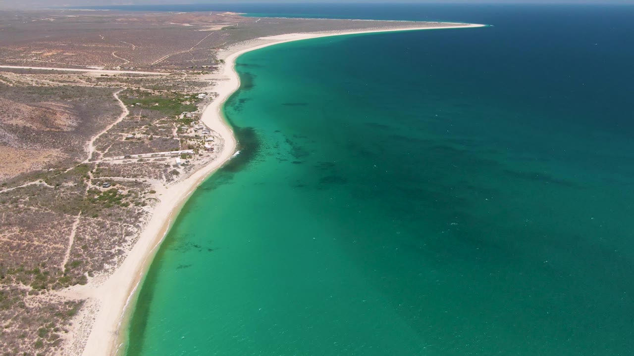 At 0:03, Playa Miramar's pale beach runs diagonally through frame, bordered by turquoise shallows, dry hills, inland tracks, and no visible people.