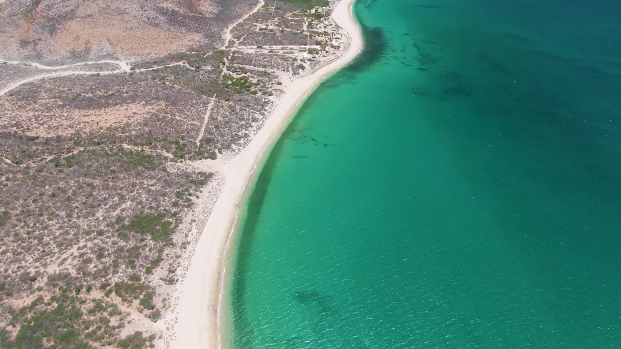 Opening aerial frame over Playa Miramar Los Cabos, with a pale beach curving between turquoise water and rocky scrubland, no visible people.