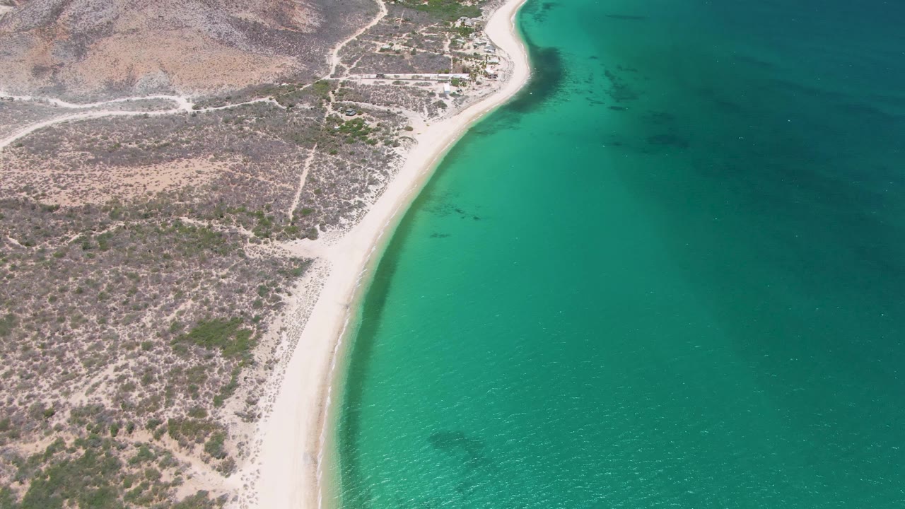 Aerial frame at Playa Miramar Los Cabos showing the shoreline slightly farther along the curve, with clear green-blue water and dry rocky ground inland, no visible people.