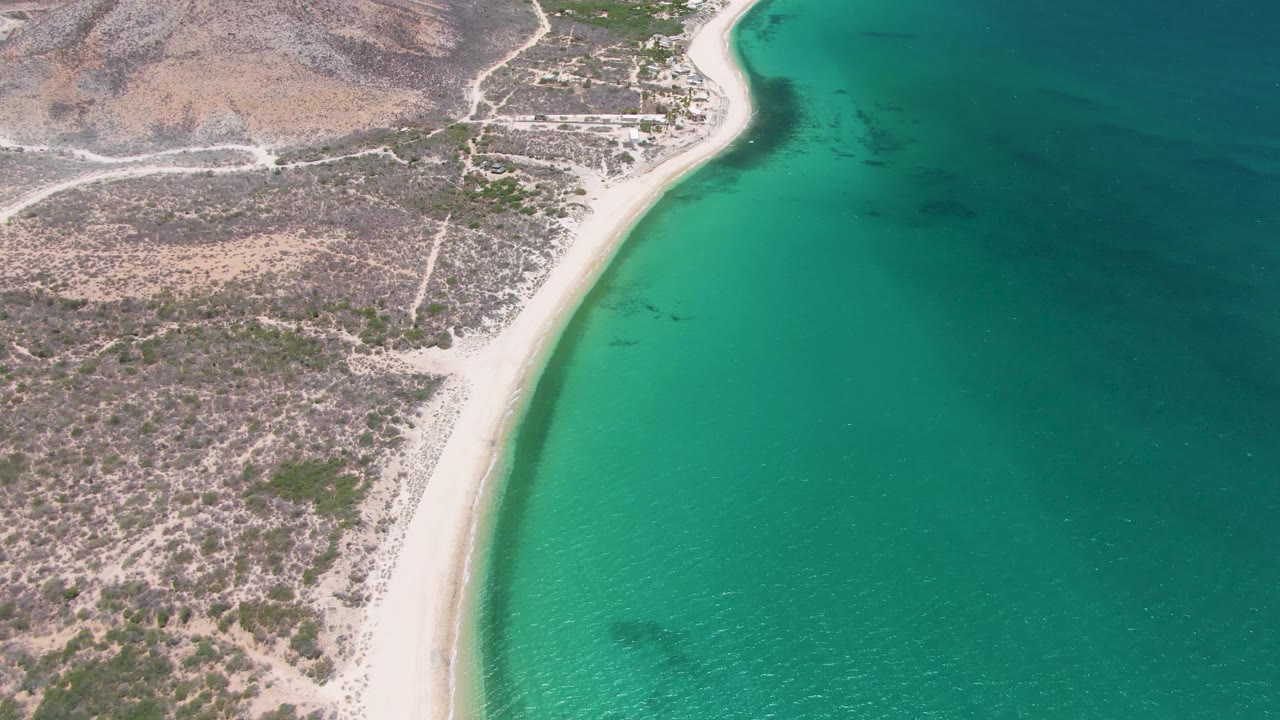 Aerial view over Playa Miramar Los Cabos with the beach edge bright against turquoise sea, inland roads and sparse scrub visible across the rocky terrain, no visible people.