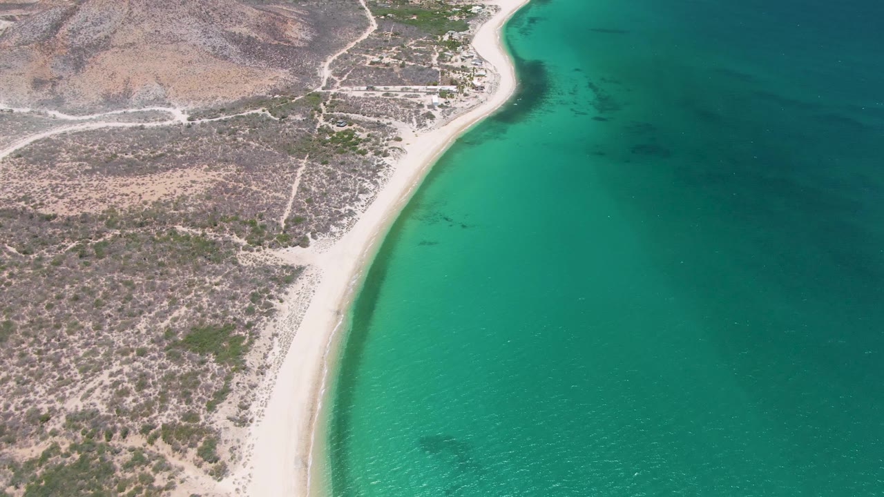 Aerial coastline with sandy curve, turquoise shallows, and arid land.