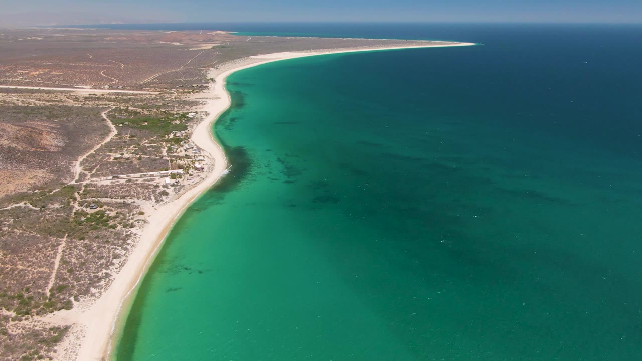 Playa Miramar Los Cabos coastline seen from above, with the sandy shore slightly farther along frame, green-blue shallows near the beach, deep water offshore, and no visible people.