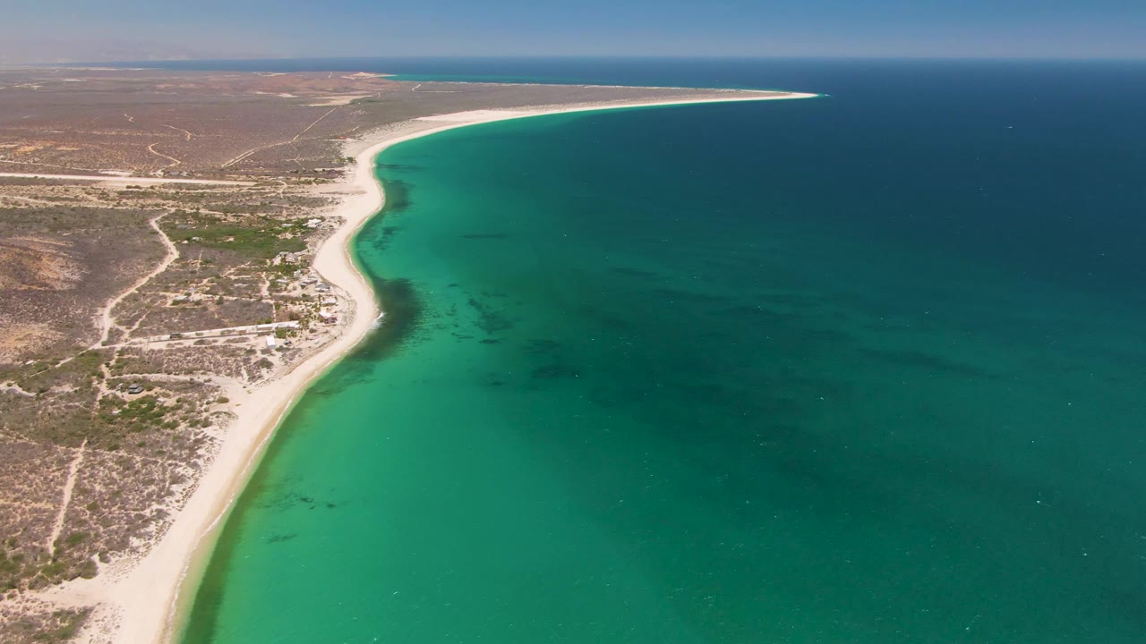 Aerial view along Playa Miramar Los Cabos shoreline, with the beach running diagonally beside green turquoise water, scrubby land to the left, open sea to the right, and no visible people.