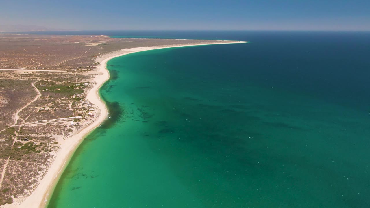 Drone view of Playa Miramar shoreline with sandy beach along dry Baja California Sur terrain and a broad band of turquoise water fading into deeper blue, no visible people.