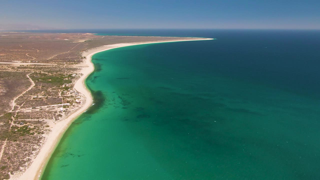 Wide aerial frame over Playa Miramar, showing the beach bending toward a narrow point in the distance with turquoise shallows, darker offshore water, and dry land at left, no visible people.
