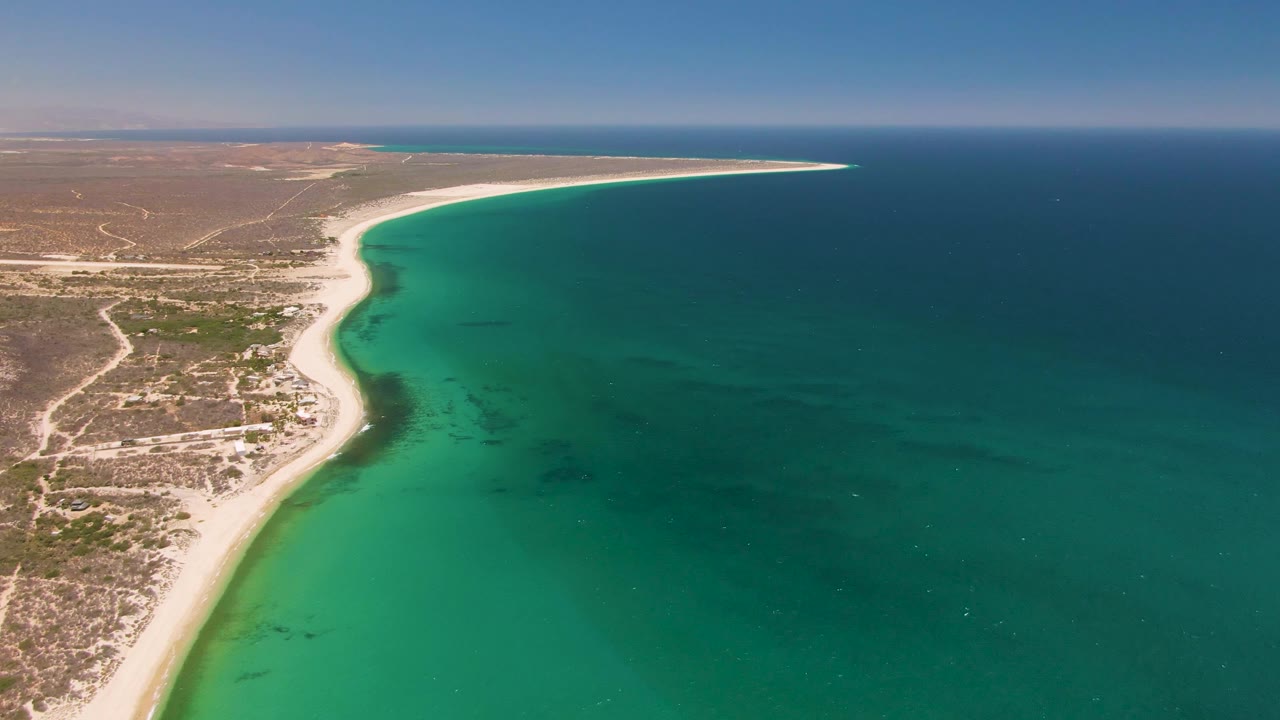Aerial view of Playa Miramar with the beach running diagonally below dry scrubland, visible dirt tracks inland, and green-blue ocean filling the right side, no visible people.