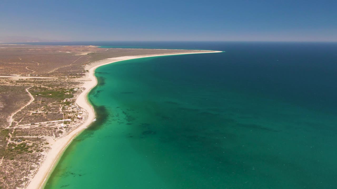 Aerial frame over Playa Miramar Los Cabos showing a pale curved beach between scrubby arid land and turquoise ocean, with no visible people.