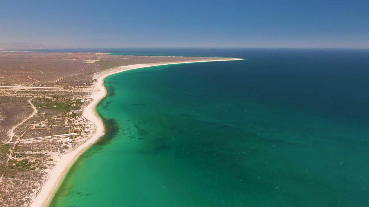 Aerial view at Playa Miramar Los Cabos with the sandy shoreline slightly more centered, dry land tracks at left, turquoise shallows, and no visible people.
