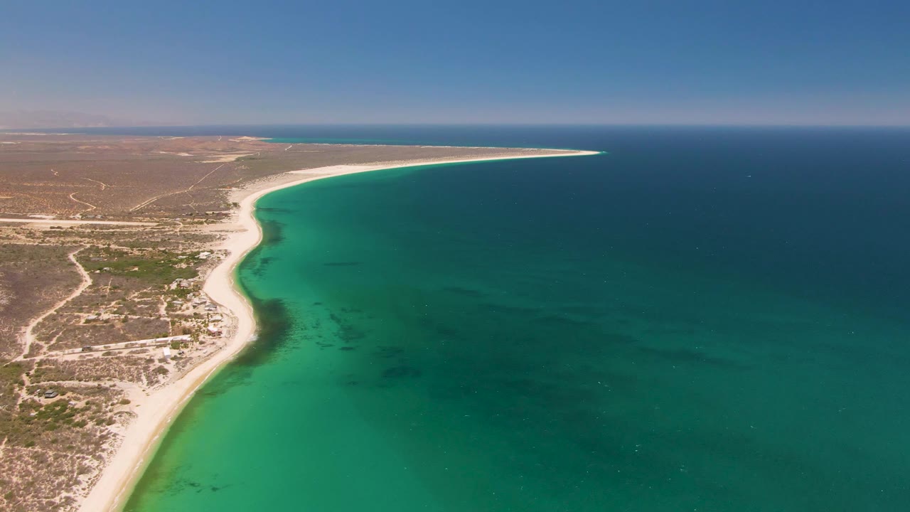 Aerial frame from Playa Miramar Los Cabos showing turquoise water filling more of the foreground, a narrow sand strip at left, arid coast beyond, and no visible people.