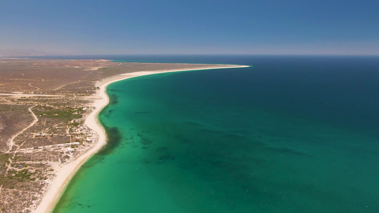 Coastal aerial with sandy beach curving through arid terrain beside clear ocean.