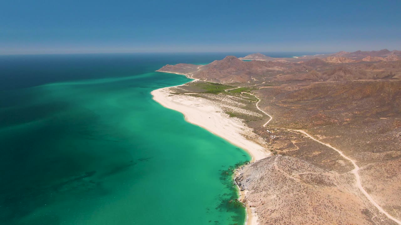 Aerial frame over Playa Miramar Los Cabos showing turquoise water, a long white sand beach, rocky desert terrain, distant mountains, and no visible people.