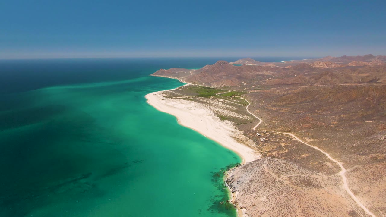 High aerial view at Playa Miramar Los Cabos with the curved beach centered, turquoise water spreading across the foreground, a dirt road inland, and no visible people.