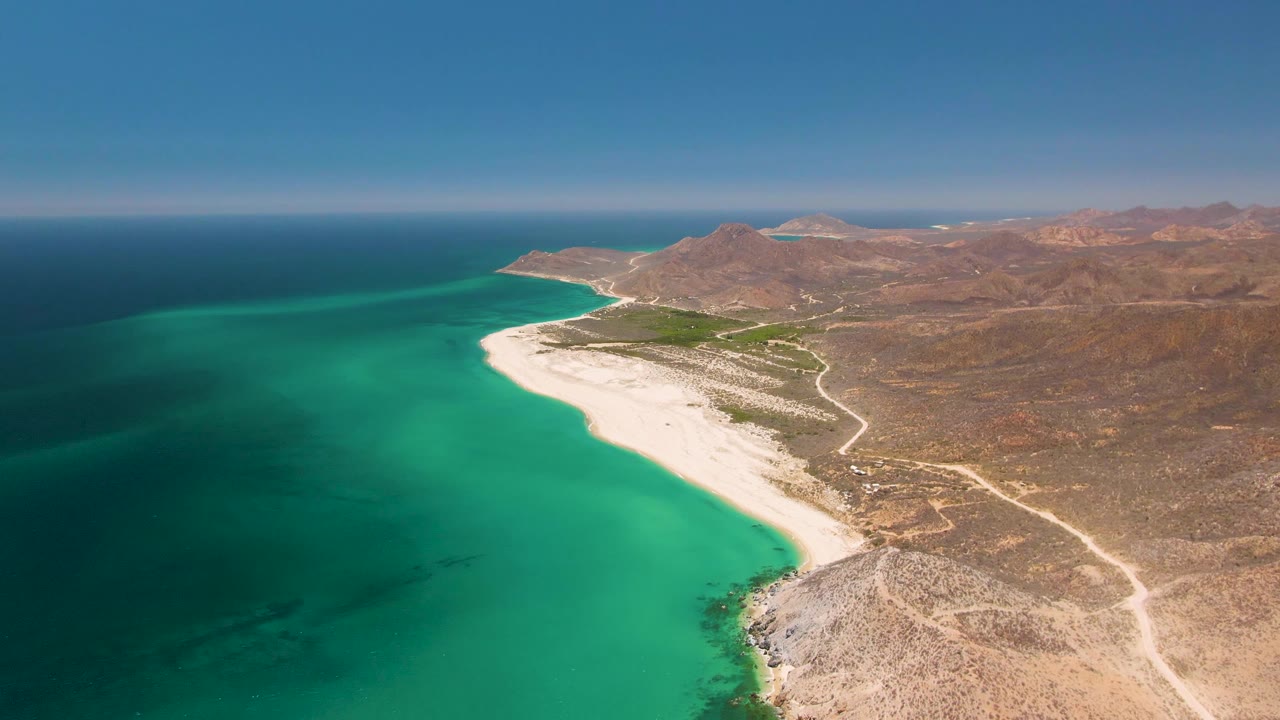 Wide drone-like frame of Playa Miramar Los Cabos with turquoise shallows, pale sand, sparse arid ground, distant ridges, a level horizon, and no visible people.