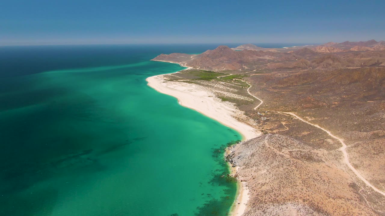 Aerial view of turquoise ocean, white beach, arid terrain, and distant mountains.