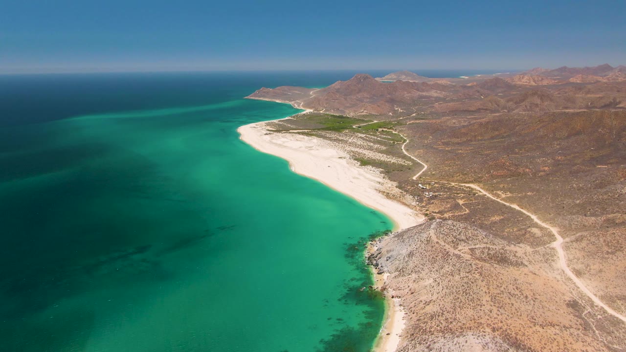 Second aerial frame at Playa Miramar Los Cabos shows a broad turquoise bay, pale beach sand, scrubby desert ground, winding dirt roads, low mountains, and no visible people.