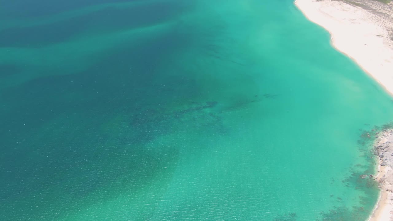 Aerial view of turquoise water beside pale sandy and rocky shoreline.